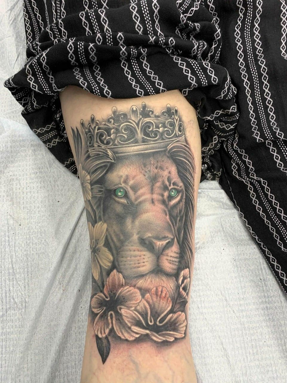A realistic crowned lion with green eyes sits above flowers in an arm tattoo.