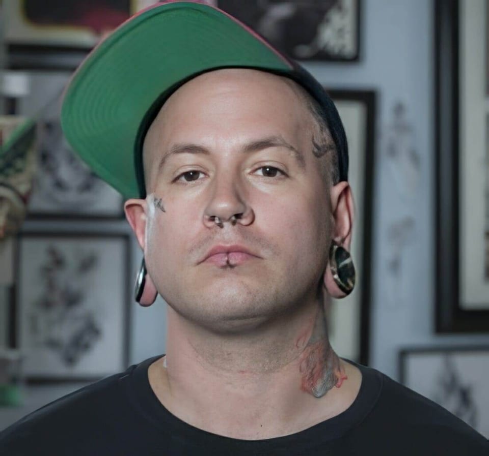 Arty faces the camera in a tilted green-brimmed cap before framed tattoo art.
