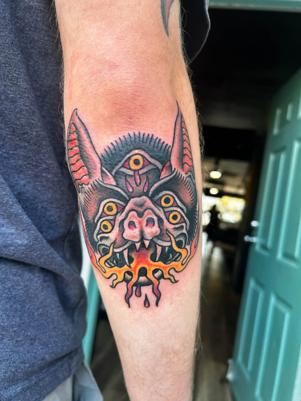 A traditional bat head with yellow eyes breathes orange flames on a forearm.