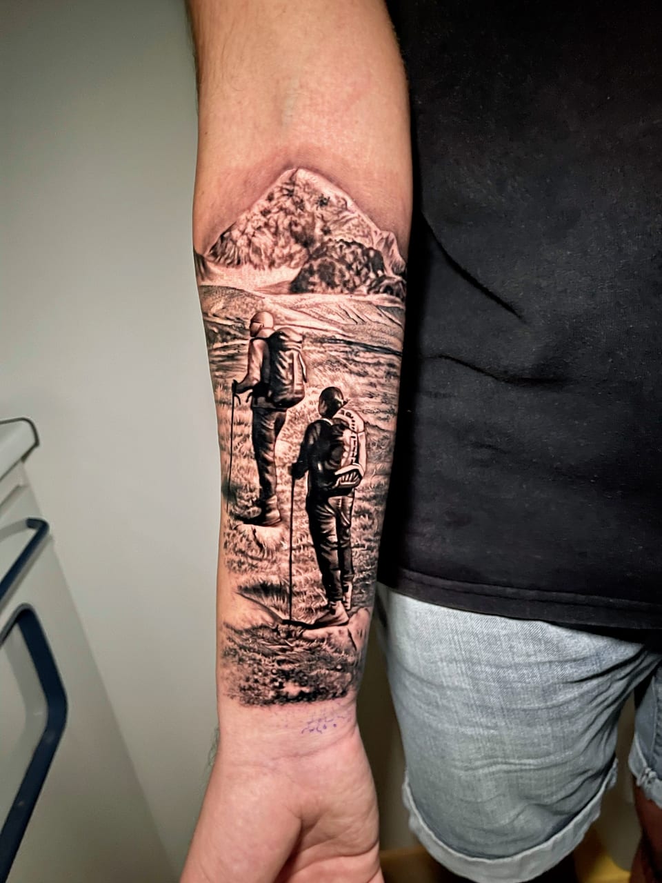 A black and grey realism hiking scene with backpackers and mountains covers a forearm.