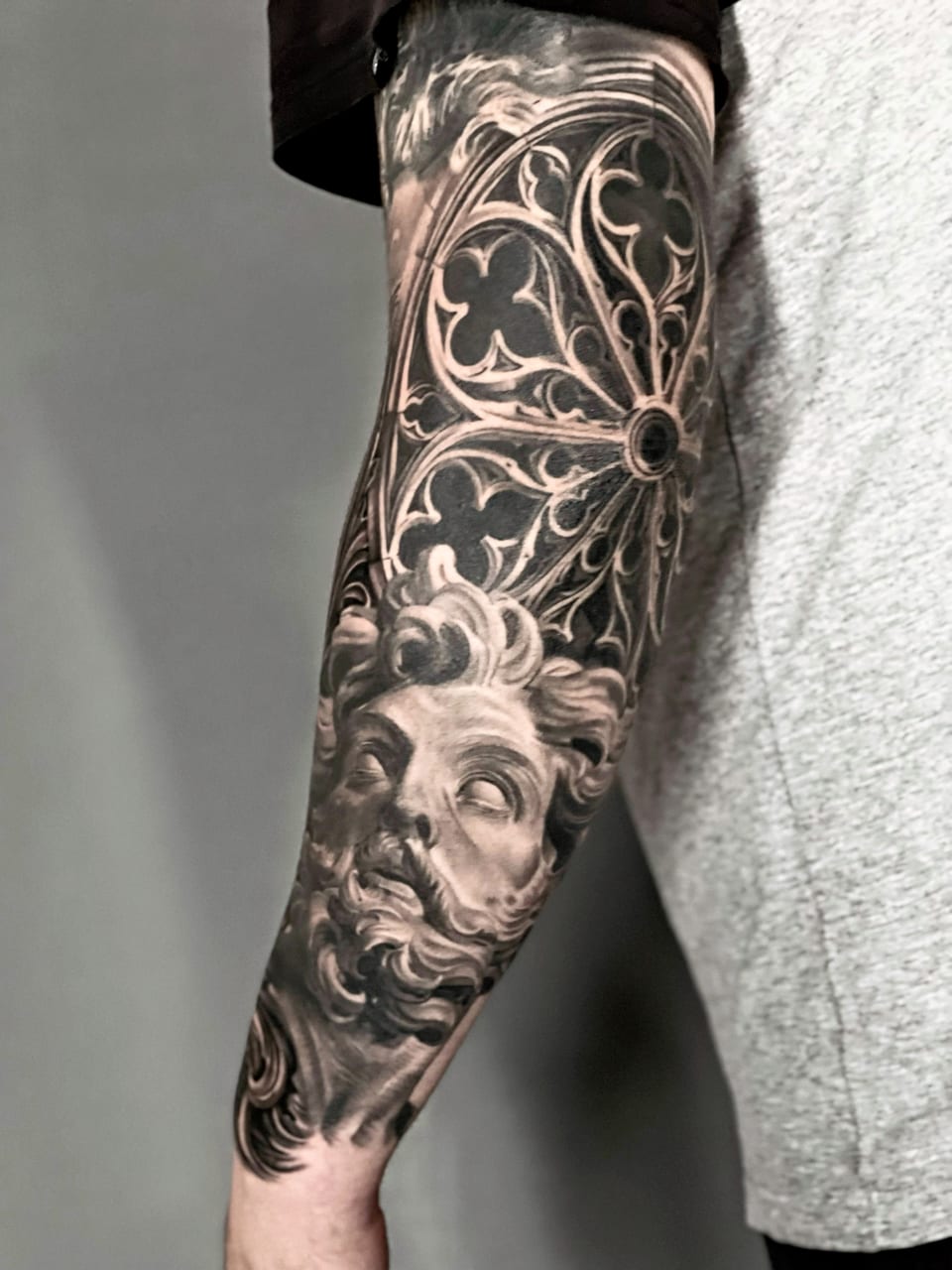 A black and grey realism bearded statue sits beneath an ornate circular window on a forearm.