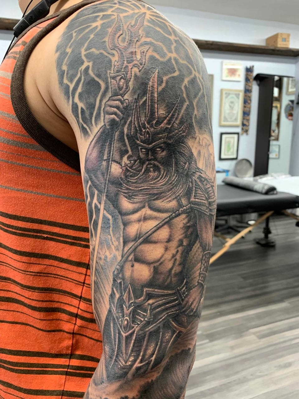 Black and grey realism tattoo of a bearded warrior with a trident and lightning covers the upper arm.