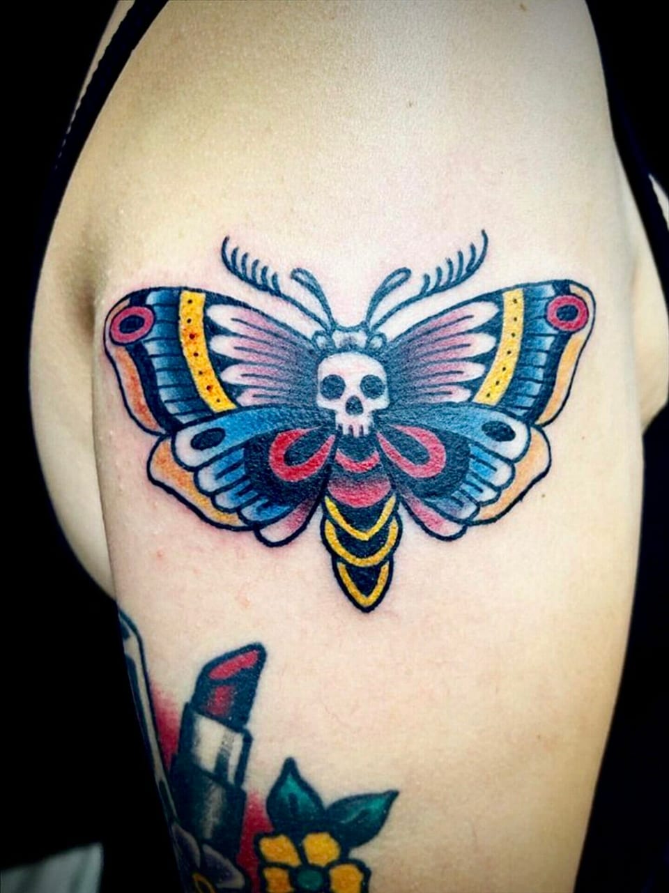 Traditional color moth with a skull on its body and patterned wings sits on the upper arm.
