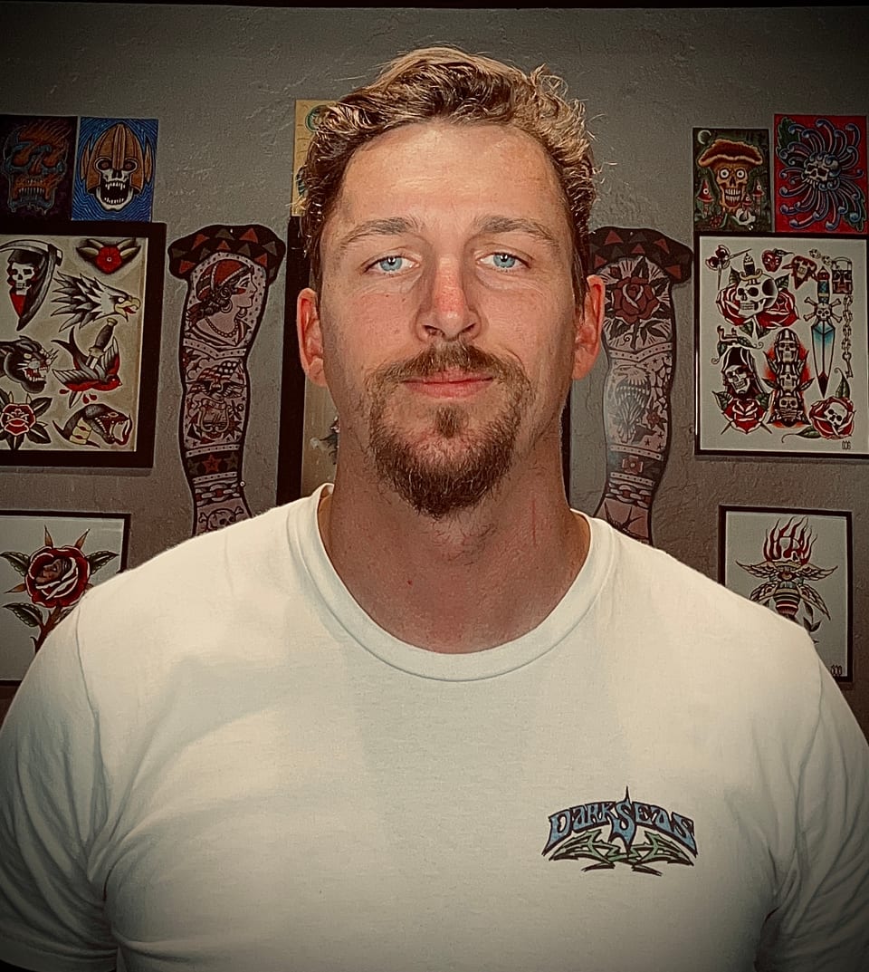 Slaine faces the camera in a white shirt in front of framed tattoo flash.