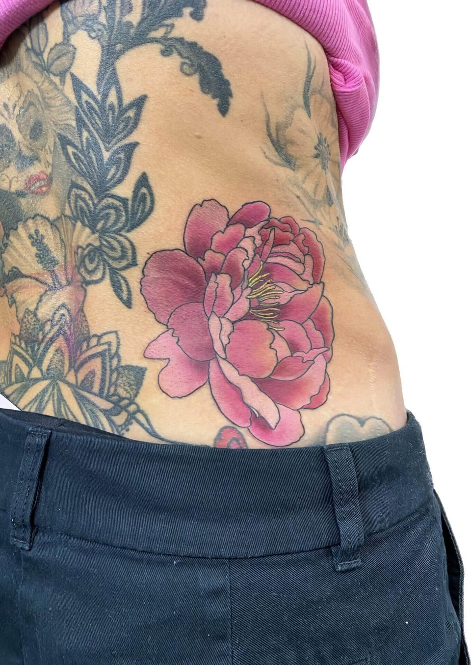 A pink illustrative peony with yellow stamens sits among floral tattoos on the lower back.