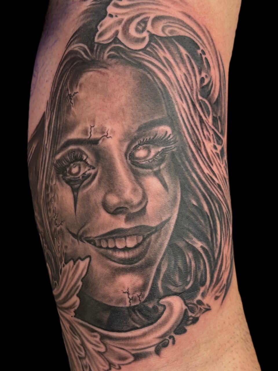 A black and grey realistic woman with clown makeup, cracked skin and a wide grin covers the forearm.