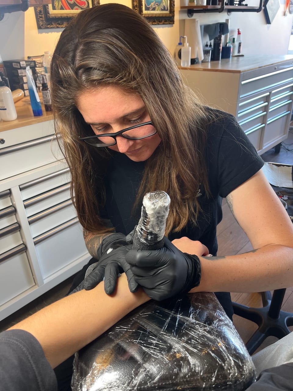 Madison Sweet wears glasses and black gloves while tattooing a client's forearm beside studio drawers.