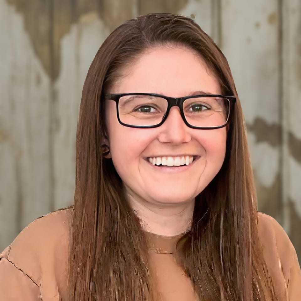Madison Sweet smiles in dark-framed glasses and a tan top against a weathered wood wall.